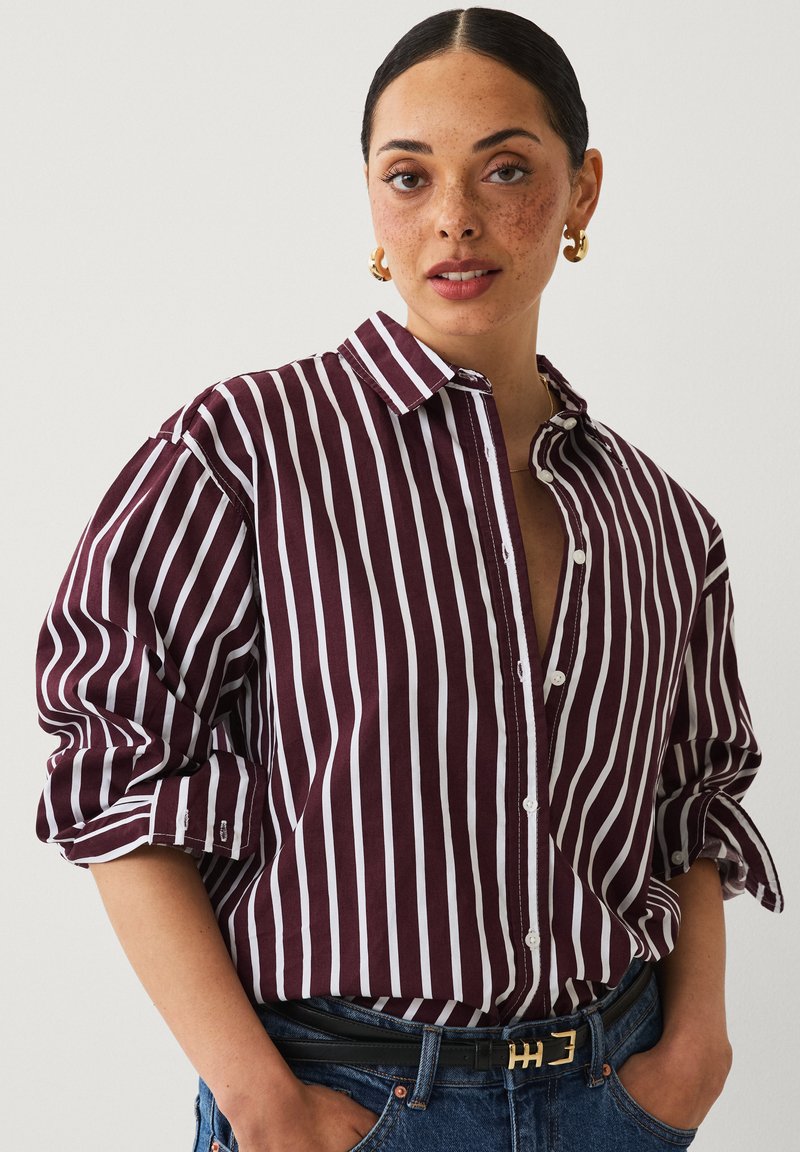 Striped shirt in dark burgundy and white, featuring a button-up front, collar, and rolled sleeves. Paired with high-waisted denim and a belt.