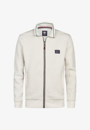 Valkoinen vetoketjullinen takki, jossa on teksturoitu kangas, sisältää pystykauluksen, jossa on vihreitä ja sinisiä raitoja, sekä rintapatch-logon.