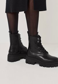 Melna ādas potīšu zābaki ar masīvu zoli, ar šņorēšanas dizainu, sānu rāvējslēdzēju un gandrīz nemanāmām šuvēm.