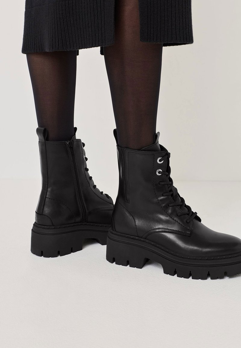 Melna ādas potīšu zābaki ar masīvu zoli, ar šņorēšanas dizainu, sānu rāvējslēdzēju un gandrīz nemanāmām šuvēm.