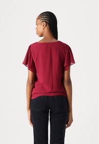 Maroon short-sleeve blouse with flutter sleeves, smooth texture, and a squared back neckline, paired with dark pants.