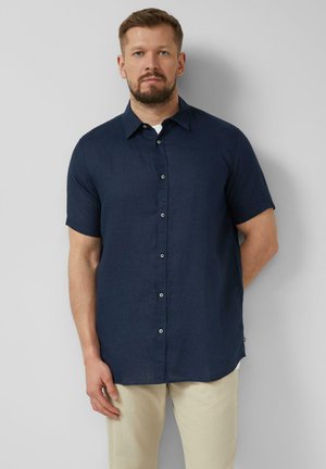 Mann mit Bart, der ein marineblaues kurzärmeliges Hemd mit Knöpfen und beige Hose trägt, steht vor einer schlichten hellgrauen Wand.