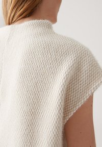 Close-up van een persoon die een gestructureerde, mouwloze crèmekleurige gebreide sweater draagt, waarbij de achterkant en een deel van de schouder zichtbaar zijn.