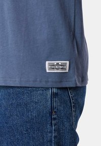 Blue cotton t-shirt with a straight cut, featuring a rectangular white label reading "DENIM FACTORY" sewn at the hem, paired with blue jeans.
