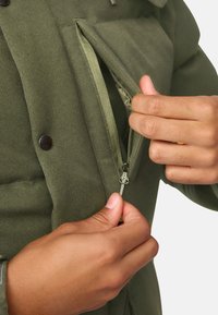 Olivengrønt overtøj med et tekstureret stof, der har en lynlåslomme. Hænder griber om lynlåsen, der fremhæver dens metaltræk og syninger.