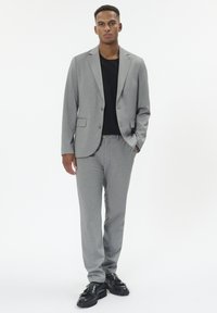 Vyras stovi dėvėdamas pilką kostiumą, juodą marškinį ir juodus elegantiškus batus prieš paprastą baltą foną.