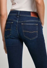 Kvinna som bär mörkblå denimjeans med sydda bakfickor och en brun läderlogga på midjebandet.