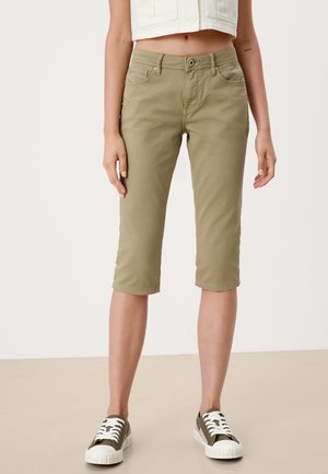 Femme portant un pantalon court kaki avec poches avant et des baskets vert olive à lacets blancs, debout devant un fond uni.