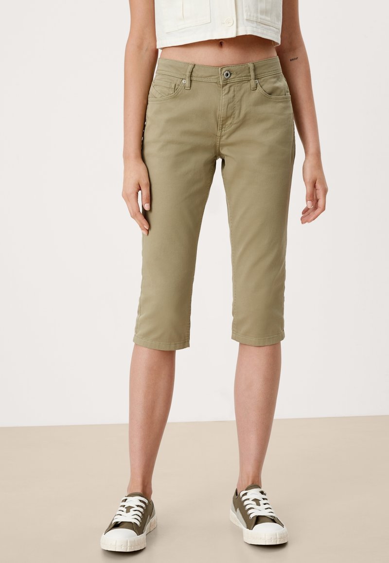 Femme portant un pantalon court kaki avec poches avant et des baskets vert olive à lacets blancs, debout devant un fond uni.