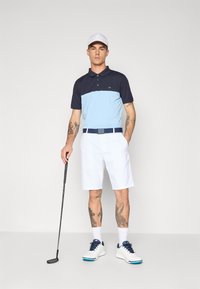 Férfi golf öltözet, amely egy sötétkék és világoskék pólóból, fehér sortból és fehér golfcipőből áll, navy kiegészítésekkel, miközben egy golfütőt tart.