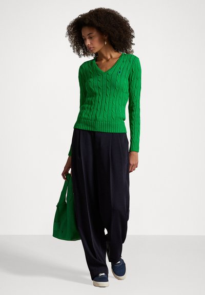 Green cable-knit sweater with a V-neck design, paired with loose black trousers and green handbag. Model wears blue slip-on shoes.