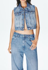Skrajšan denim jopič v svetlo modrem pranju, z gumbasto zaponko, dvema žepoma na prsih in raztrgano obrobo, v kombinaciji z modrimi hlačami visoke pasu.
