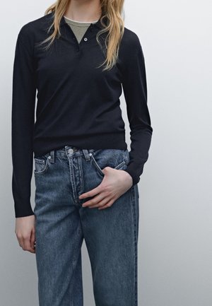 Person wearing a black long-sleeve top over a green shirt and blue high-waisted jeans, with one hand in a pocket against a plain background.