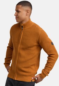 Pomarańczowy sweter na zamek błyskawiczny z wysokim kołnierzem, teksturowana tkanina, ściągacze na mankietach oraz mała czarna łatka z logo na rękawie.