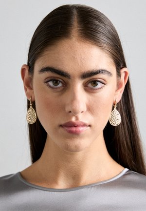 Gold teardrop earrings with intricate cut-out design, hung from a small hoop, worn by a model with straight, dark hair and a gray top.