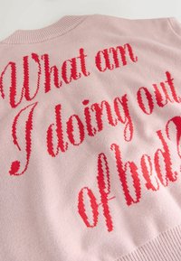 Světle růžový svetr s červeným textem vyšitým kurzivou: "Co tu dělám mimo postel?" Pletená textura s žebrovaným límcem.
