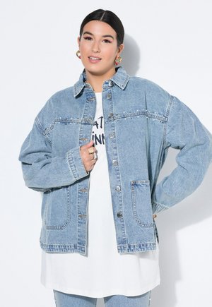 Tummatukkainen nainen, joka käyttää kultaisia rengaskorvakoruja, ylisuurta vaaleansinistä niititettyä farkkutakkia, valkoista graafista paitaa ja vaaleita farkkuja.