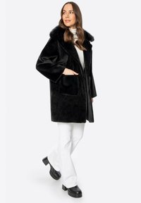 Abrigo de piel sintética negro con un cuello ancho, dos bolsillos delanteros y un corte holgado; combinado con pantalones acampanados blancos y botas de plataforma negras gruesas.