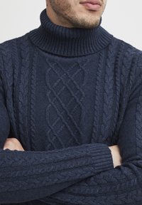 Homme portant un pull à col roulé en tricot bleu marine, bras croisés et barbe de trois jours visible, sur un fond uni.