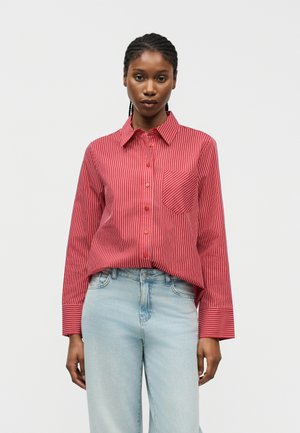 Γυναίκα που φοράει κόκκινο πουκάμισο με άσπρες κάθετες ρίγες και ανοιχτόχρωμο τζιν ψηλόμεσο, στέκεται μπροστά σε απλό φόντο.