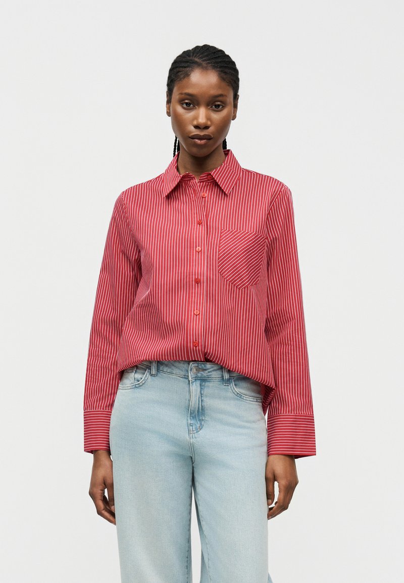 Ženska, oblečena v rdečo srajco z belimi navpičnimi črtami in svetlo modre jeans hlače z visokim pasom, stoji ob enobarvnem ozadju.