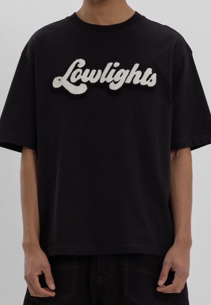 Personne portant un t-shirt noir à manches courtes avec l'inscription « Lowlights » en lettres blanches texturées sur la poitrine.