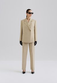 Costume oversized à carreaux beige avec col châle, fermeture à un bouton, pantalons plissés, gants noirs et lunettes de soleil noires angulaires.