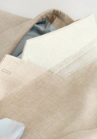 Blazer de lino beige con un suave forro azul claro. El bolsillo interior presenta un borde de tela crema suave con detalles de costura precisos.