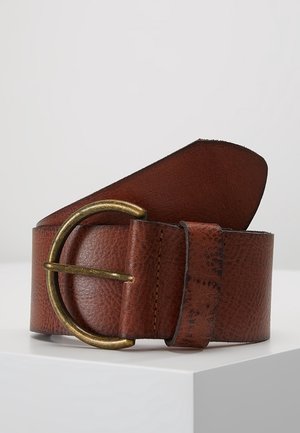 Ceinture en cuir marron roulée avec une grande boucle ronde en laiton sur une surface blanche avec un fond gris.