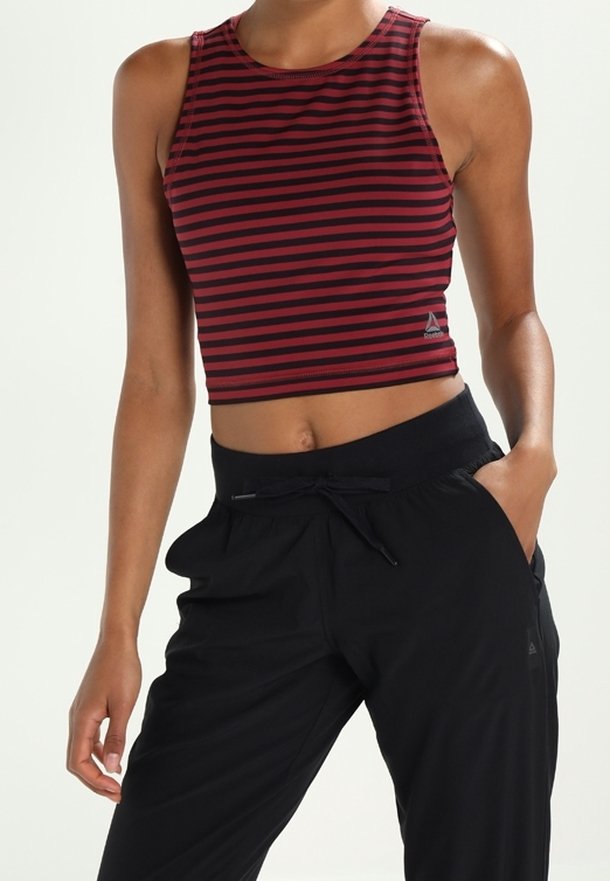 Femme portant un crop top sans manches rayé rouge et noir horizontalement et un pantalon noir à cordon, debout avec une main dans la poche.