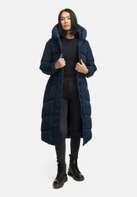 Mörkblå lång pufferjacka med huva, försedd med quiltade sektioner och dragkedja framtill. Kombinerad med svarta jeans och kraftiga svarta stövlar.