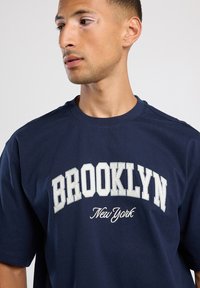 Tummansininen puuvillapaita, jossa on pyöreä kaula-aukko. "BROOKLYN" ja "New York" on painettu valkoisella, ja siinä on rohkea serif-fontti. Lyhyet hihat.