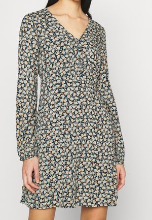 Kobieta ubrana w sukienkę z długim rękawem, z dekoltem w serek, w drobny wzór kwiatowy w kolorach niebieskim, beżowym i czarnym.