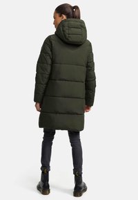 Padded long coat in dark green with a hood, featuring a quilted design and ribbed texture. Worn with dark jeans and sturdy black boots.
