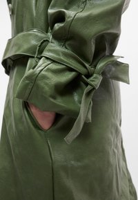 Πράσινο δερμάτινο παλτό με ζώνη στη μέση και πλαϊνή τσέπη. Η υφή είναι λεία με ελαφριά γυαλάδα, υπογραμμίζοντας τον ραμμένο σχεδιασμό του.