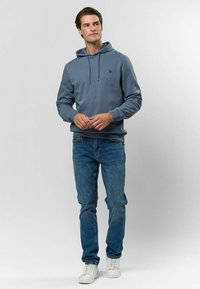 Mikina s kapucňou z mäkkej, tlmenej modrej látky s nenápadným logom na hrudi, v kombinácii so svetlomodrými džínmi a bielymi teniskami.