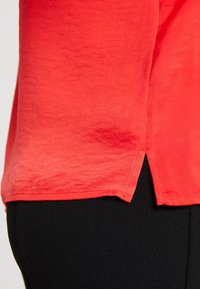 Top rosso a maniche corte realizzato in tessuto morbido, con orlo leggermente arrotondato e spacchi laterali, abbinato a pantaloni neri con texture.
