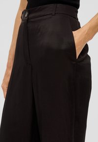 Černé, lehké kalhoty s hladkou texturou, se boční kapsou a jedním knoflíkem na zapínání v pase. Minimalistický design.