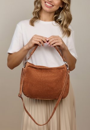 Usmievajúca sa žena v bielom topu a béžovej sukni drží hnedú semišovú kabelku s dvoma popruhmi na neutrálnom pozadí.