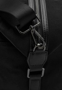 Bolso negro con un material texturizado, una correa ancha y un clip de metal. La cremallera que recorre el lado añade un detalle funcional.