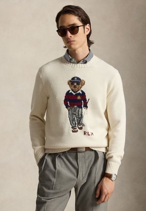 Mann mit Sonnenbrille, cremefarbenem Pullover mit einem Teddybär in einem marineblau-rot gestreiften Rugbyshirt, grauen Hosen und Armbanduhr.