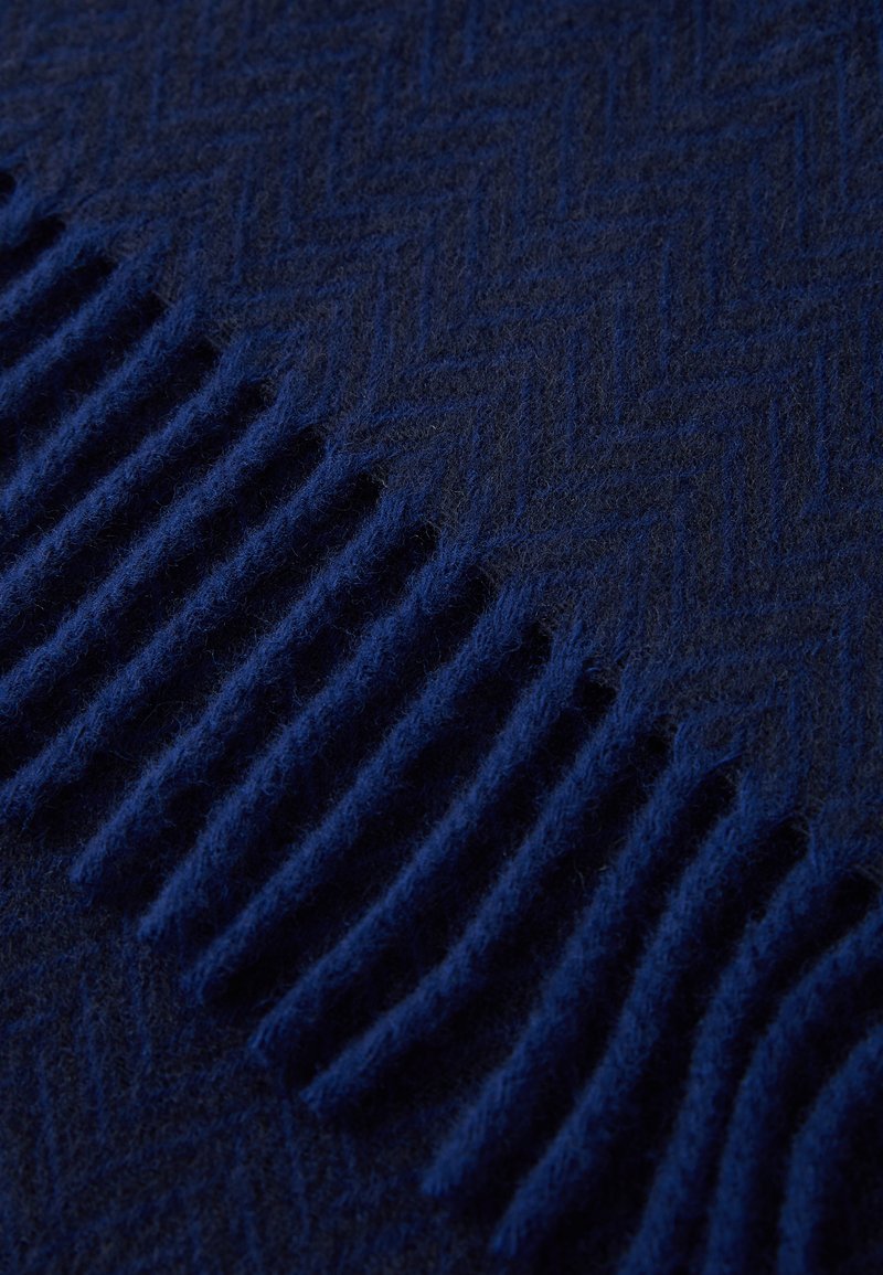 Couverture en laine de couleur bleu foncé avec un motif en chevrons et des franges texturées d'un côté. Texture douce et chaude avec une couleur uniforme.