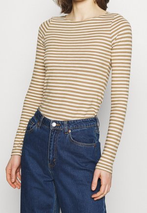 Osoba ubrana w beżowo-białą koszulę w paski z długim rękawem, włożoną do wysokich, niebieskich dżinsów, z rękami swobodnie opuszczonymi wzdłuż ciała.