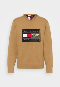 Maglione color cammello in tessuto strutturato, caratterizzato da una toppa rettangolare nera con mappa del mondo, dettagli rossi e bianchi e polsini a coste.