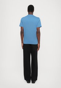 Modra majica s kratkimi rokavi in okroglim izrezom, gladke teksture in prileganja, v kombinaciji s črnimi širokimi hlačami in črnimi čevlji.