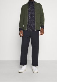 Grön zip-up jacka över en marinblå blommig skjorta, i kombination med marinblå byxor och vita sneakers. Materialen verkar mjuka med en ledig passform.