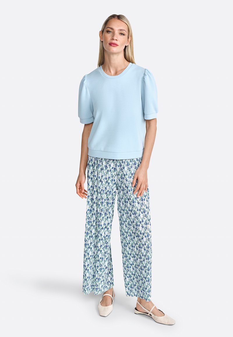 Haut bleu clair à manches courtes avec des manches bouffantes et un col rond, associé à un pantalon floral à jambes larges dans des teintes blanches et bleues. Des ballerines beiges.