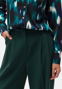Blusa multicolore e a fantasia con tonalità di blu e nero, abbinata a pantaloni verdi a vita alta con pieghe e una texture di tessuto liscia.