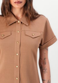 Kortsleppet skjorte med krage i lys brun, ribbet stoff. Har to frontlommer og detaljer med gullknapper.