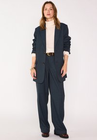 Completo blu scuro a righe con una silhouette rilassata; presenta una giacca doppiopetto e pantaloni a gamba larga, abbinati a un dolcevita crema.